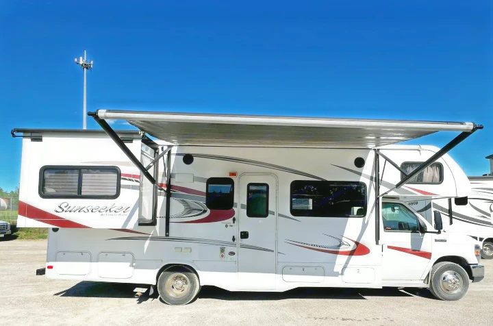 Side view of a white Sunseeker RV rental with red/gray graphics and an extended awning under a clear blue sky.. Forest River Sunseeker 2016