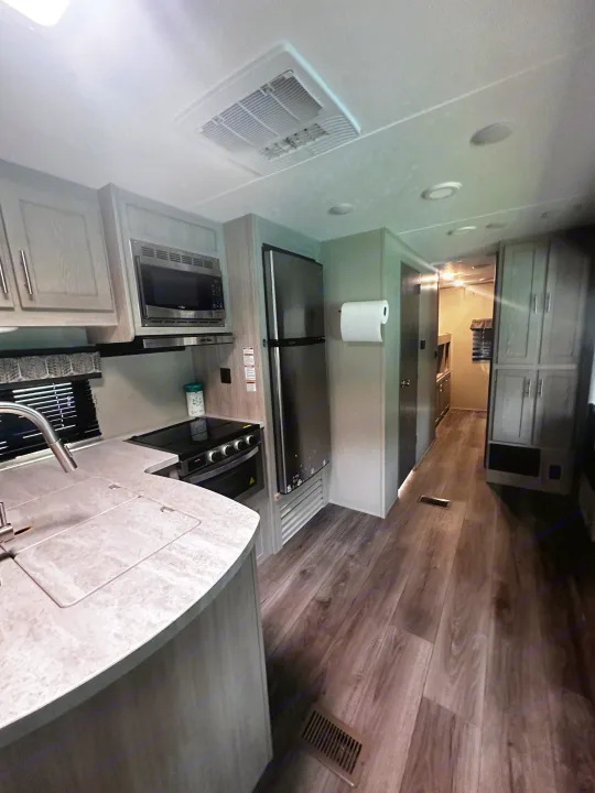 Interior of an RV rental, featuring a gray-toned kitchen with sink, microwave, fridge, and a hallway with wood-look flooring.. Coachmen 343bhts 2023