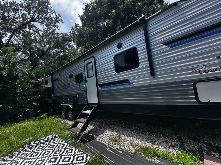 Side view of a travel trailer, a great RV rental. Features open door with steps, mat on grass, gravel ground.. Coachmen 343bhts 2023