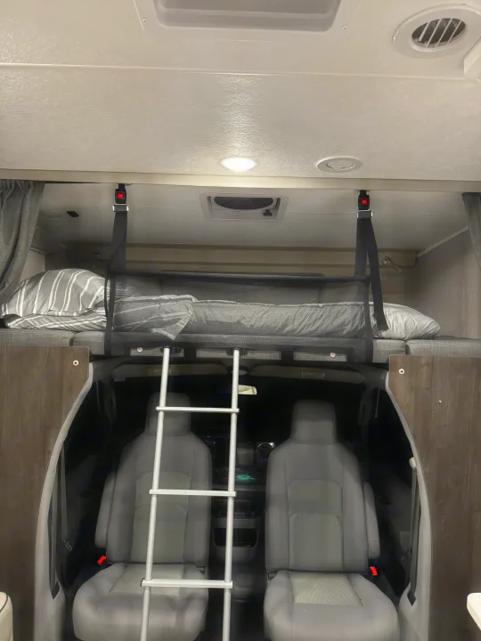 Inside an RV rental: a spacious over-cab bunk bed with safety net and ladder, viewing the front cockpit and grey seats.. Forest River Coachmen Leprechaun Premier 2022
