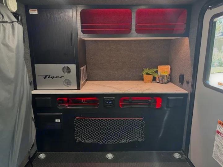 This RV rental showcases a compact interior with a "Flyer" cooler, modern countertop, and red-lit storage ideal for travel.. InTech RV Flyer 2023