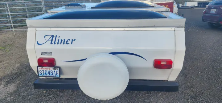 Rear view of a white Aliner pop-up camper for rv rental, displaying a Washington license plate and a rear-mounted spare tire.. A Liner A Liner Popup Trailer 2006