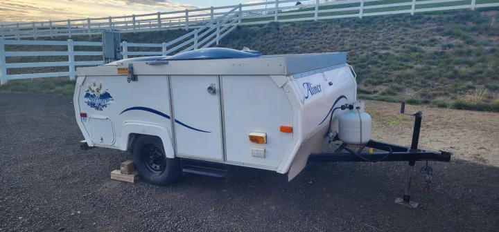 A white Aliner Sportliner pop-up RV camper parked on gravel with a fence and grassy hill behind. Perfect for your rv rental!. A Liner A Liner Popup Trailer 2006