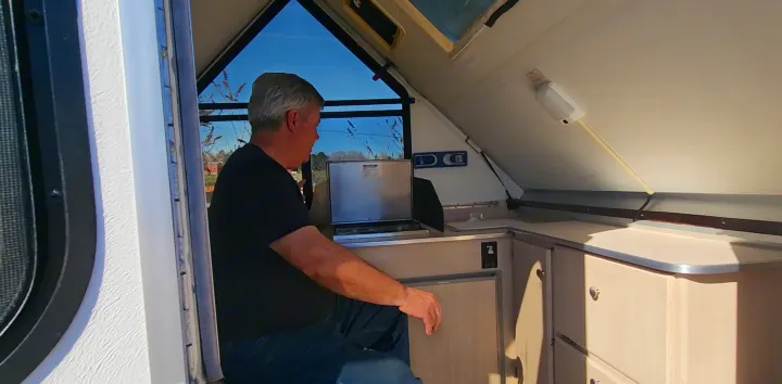 A man is seated within an RV rental A-frame camper, with large windows overlooking trees. Features a compact kitchen & fridge.. A Liner A Liner Popup Trailer 2006