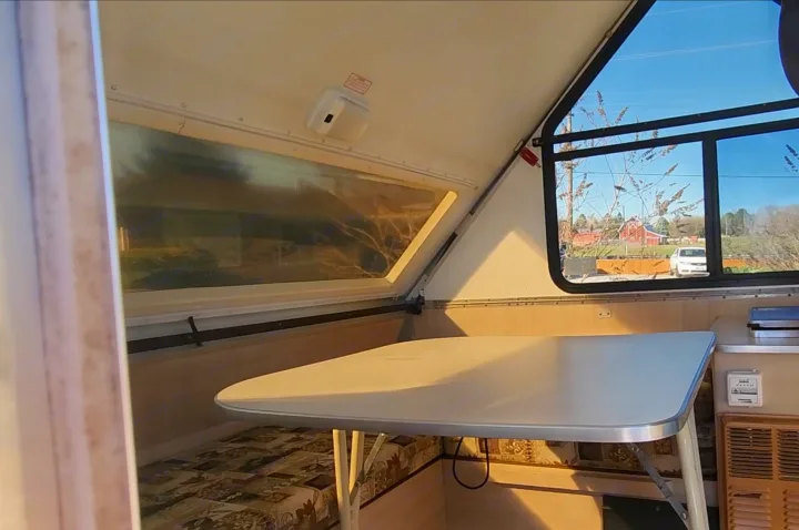 Bright A-frame rv rental interior showing a dining table, window with a red barn view, and patterned seating.. A Liner A Liner Popup Trailer 2006