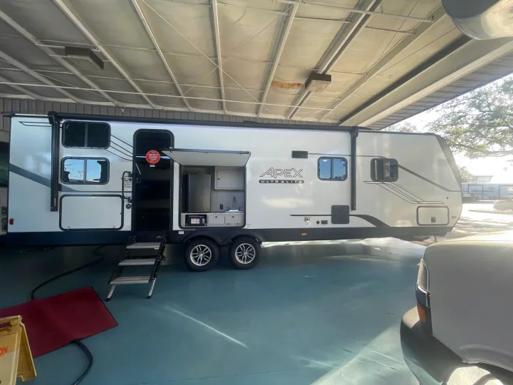 Apex Ultralite travel trailer, ideal for RV rental, showcasing its open outdoor kitchen and steps, parked inside.. Coachmen APEX 284BHSS 2025