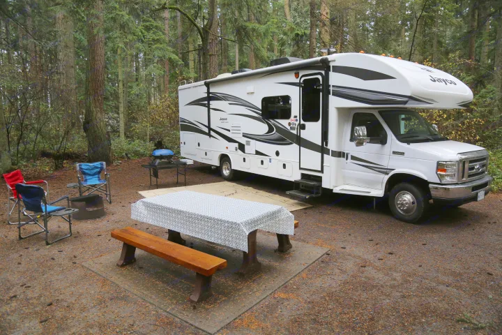 Enjoy nature with this RV rental. A Jayco motorhome is set up at a forest campsite with a picnic table, grill, and chairs.. Jayco Greyhawk 2019