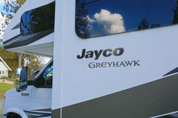 A white Jayco Greyhawk RV for rent, ready for your next adventure. Features sky and tree reflections on its sleek side.. Jayco Greyhawk 2019