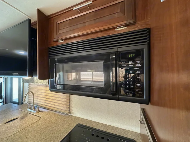 RV rental kitchen: black over-range microwave with digital display, wooden cabinets, sink, faucet, and a wall-mounted TV.. Forest River Fr3 2019