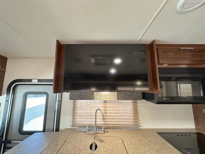 RV rental kitchen showing a TV mounted above a sink with faucet, microwave, and cooktop. Exit door on left.. Forest River Fr3 2019