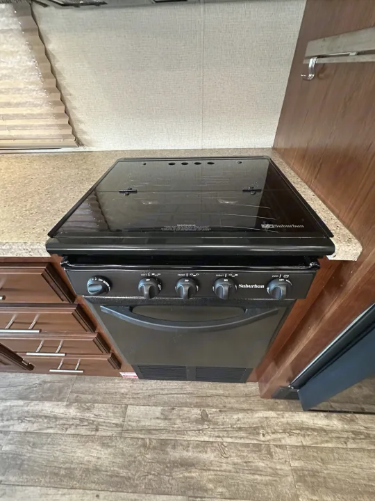 This RV rental kitchen features a black Suburban stove and oven unit with a sleek glass top, next to wooden cabinets.. Forest River Fr3 2019