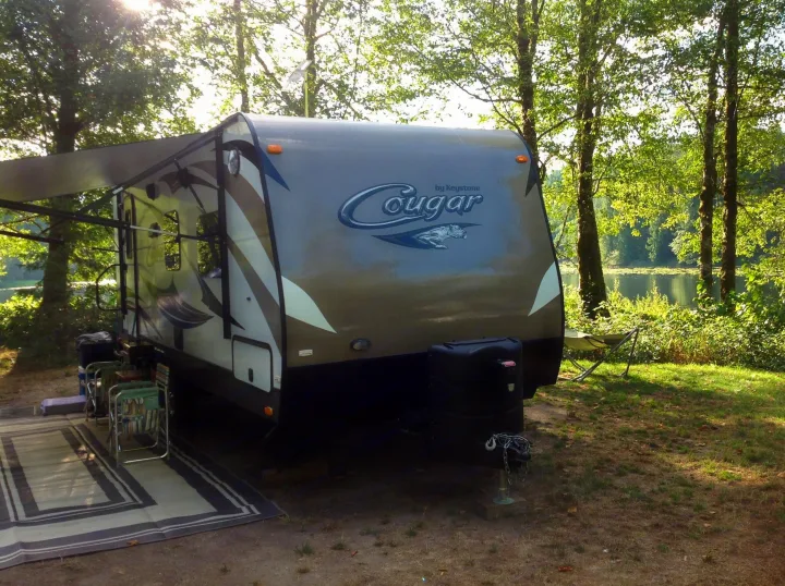 Enjoying an RV rental at a scenic lakeside campsite with a Cougar travel trailer, awning out, and outdoor seating.. Keystone RV Cougar 2015