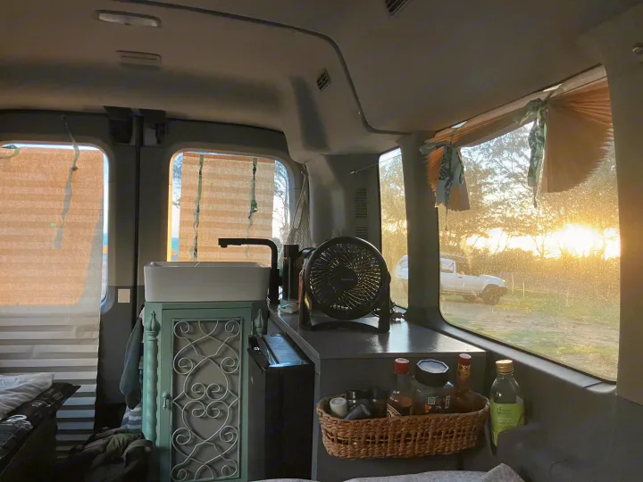 Inside an RV rental: a compact kitchen with sink, fan, and storage, overlooking a bright, tree-lined sunset view.. Ford Transit 2018