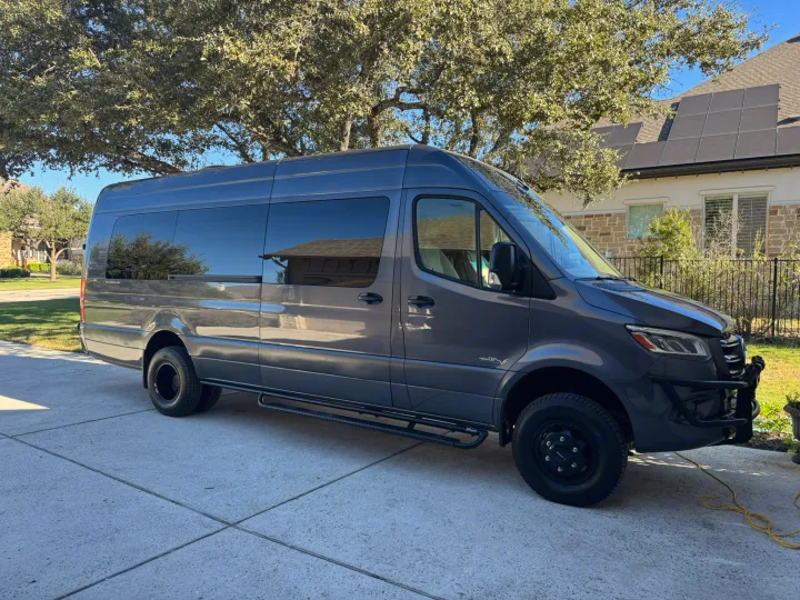 This customized dark gray Mercedes Sprinter van, ideal for rv rental adventures, is parked on a sunny driveway beside a house and trees.. Midwest Automotive Designs Ultimate Presidential 2020