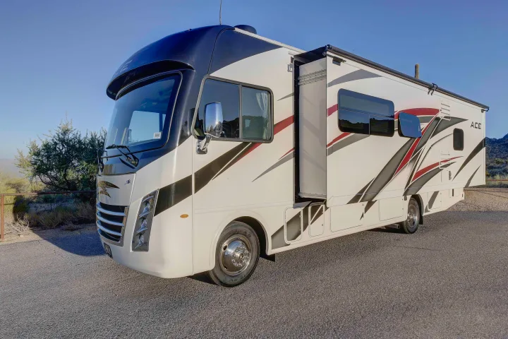 White Class A rv rental with red and black graphics, slide-out extended, parked on asphalt in a sunny desert landscape.. Thor A.C.E. 2022