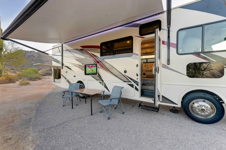 RV rental motorhome with extended awning, illuminating LED lights, an outdoor TV showing sports, plus chairs and table, set in a desert landscape.. Thor A.C.E. 2022
