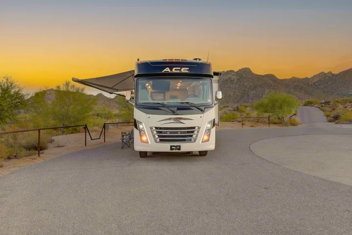 An 'A.C.E.' RV rental parked at a desert campsite with its awning extended during a vibrant sunset, surrounded by mountains.. Thor A.C.E. 2022