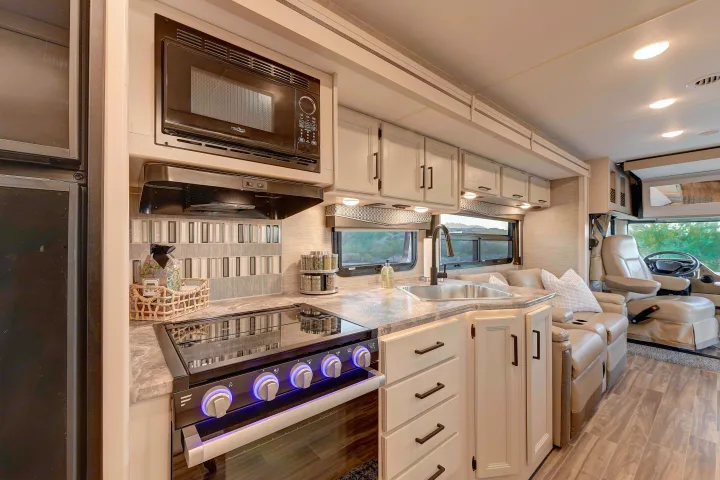 This modern RV rental kitchen features a microwave, stove with illuminated knobs, sink, and light cabinetry, leading to a comfy living area.. Thor A.C.E. 2022
