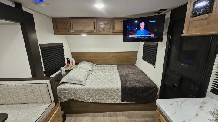 Relax in this RV rental's cozy bedroom: patterned bed, wall-mounted TV, overhead cabinets, and adjacent seating.. Forest River 17JW 2025