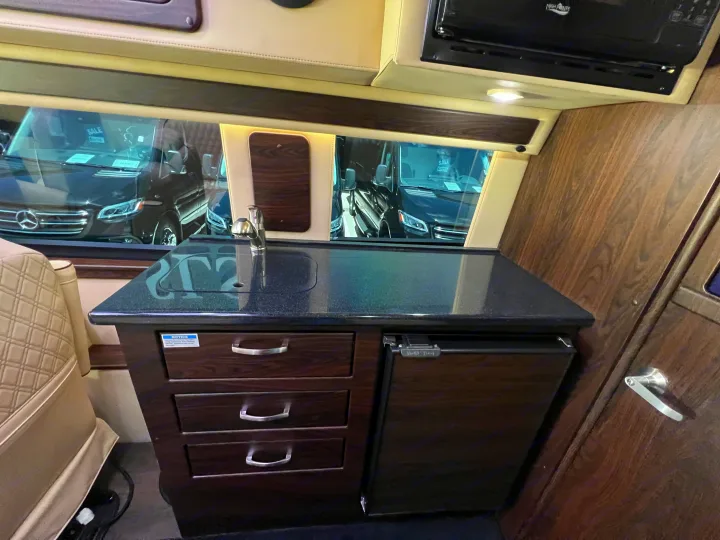 Compact RV rental kitchen area features a dark countertop, sink, faucet, microwave, and under-counter refrigerator.. Midwest Automotive Designs Day Cruiser 2024