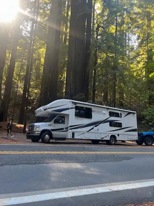 White RV rental parked among towering redwoods with sunbeams, a tow vehicle attached. Scenic forest road trip.. Entegra Coach Odyssey 2024