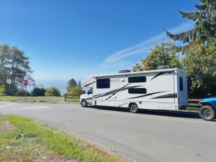Scenic RV rental with ocean view. This grey motorhome, towing a Jeep, is parked at a beautiful coastal overlook.. Entegra Coach Odyssey 2024