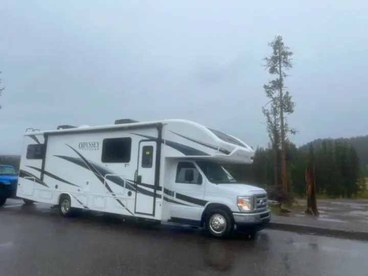 A white RV rental is parked on a wet road in a wooded setting under a cloudy sky. Ready for adventure!. Entegra Coach Odyssey 2024