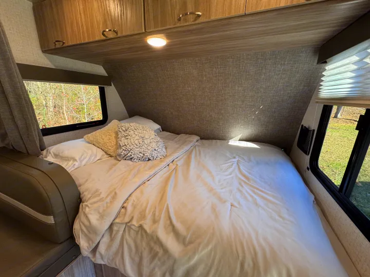 Cozy RV rental bedroom featuring a bed with white linen, overhead cabinets, and windows showing an outdoor forest view.. Winnebago Minnie Winnie 2022