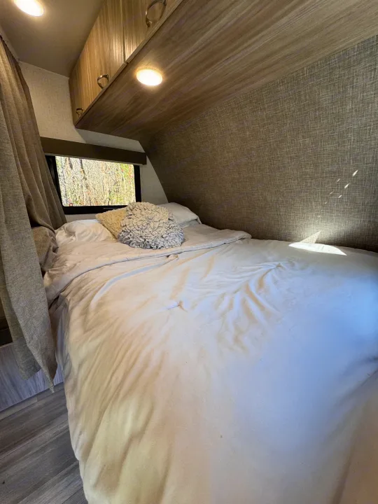 Cozy RV rental bedroom with white bedding, patterned walls, overhead cabinets, and a window view of trees.. Winnebago Minnie Winnie 2022