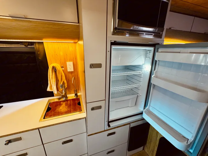 Compact RV rental interior featuring a functional kitchen with sink, microwave, and an empty, open refrigerator.. Noovo Lite 2023