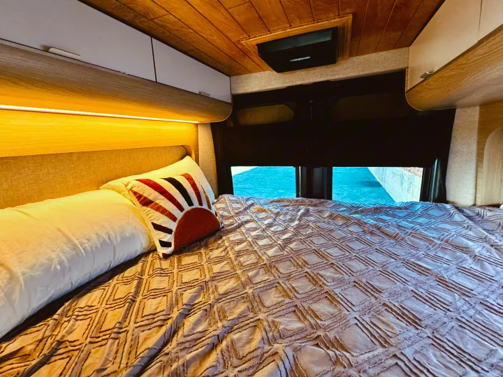 Comfortable RV rental bed featuring a textured blanket, decorative pillow, and warm overhead lighting with storage cabinets.. Noovo Lite 2023