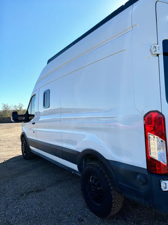 Rugged white camper van for RV rental, with all-terrain tires & roof rack, parked on gravel on a sunny day.. Ford Ford Transit 2020