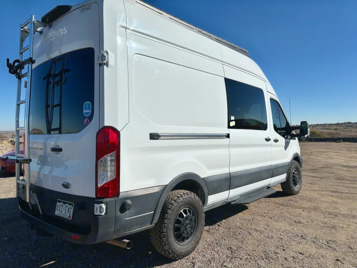 A white Ford Transit RV rental van from the rear-side view, featuring a ladder, roof rack, and rugged tires.. Ford Ford Transit 2020