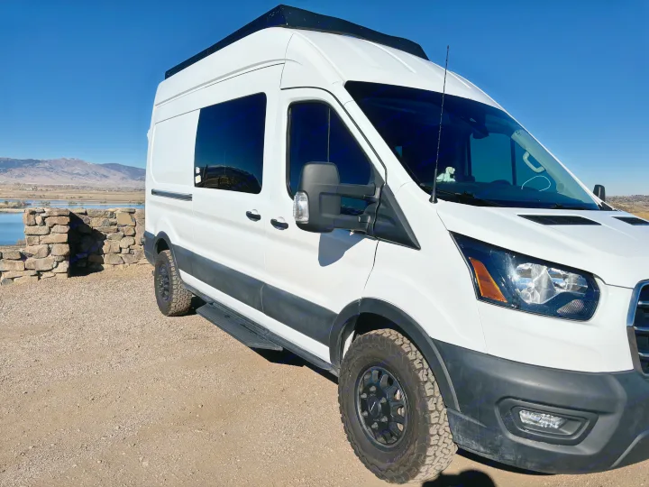 White Ford Transit camper van, an RV rental, on a gravel road with mountains and a lake in the background.. Ford Ford Transit 2020
