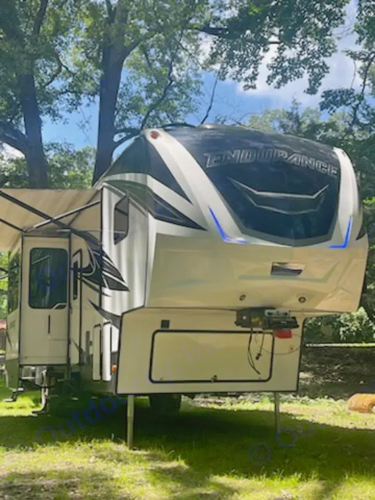 This large white "Endurance" fifth-wheel RV rental is parked on green grass under shade trees with its awning extended.. Dutchmen 3586 2019