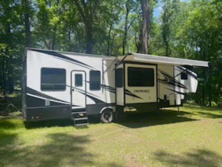 Spacious white and black fifth-wheel RV rental parked in a grassy, wooded area with its awning out, ready for camping adventures.. Dutchmen 3586 2019