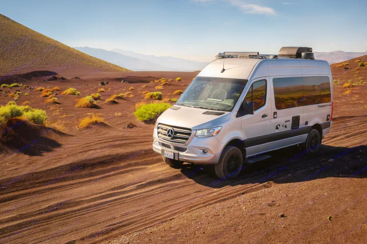 Silver Mercedes Sprinter **rv rental** van on dark volcanic sand with desert plants and distant mountains under a clear sky.. Thor Tranquility 2023