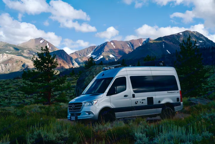 Explore mountains with a silver RV rental van. Parked amidst peaks & blue sky, ideal for your next outdoor adventure.. Thor Tranquility 2023