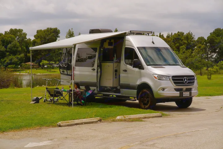 Family enjoys their silver Mercedes Sprinter rv rental, complete with an extended awning setup beside a scenic lake.. Thor Tranquility 2023