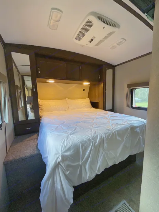 RV rental bedroom features a neatly made bed with white bedding, overhead AC, cabinets, and a window with an outdoor view.. Keystone RV Outback Super-Lite 2018