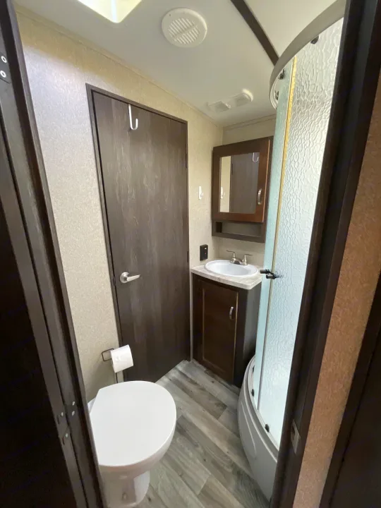 An RV rental's compact bathroom, featuring a white toilet, wood vanity with sink, and curved glass shower.. Keystone RV Outback Super-Lite 2018