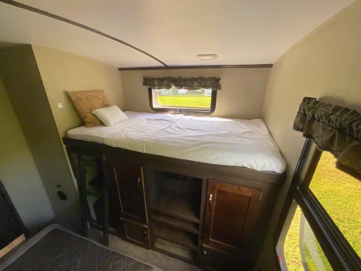 RV rental bunk bed area: elevated bed with white sheets & pillows, built-in storage, and window with outdoor view.. Keystone RV Outback Super-Lite 2018