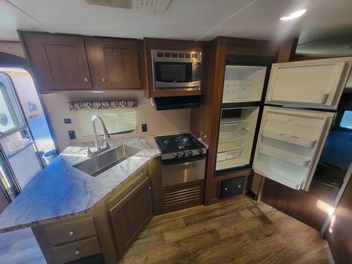 This RV rental's kitchen boasts a sink, microwave, stove, and an open refrigerator, offering modern comfort and ample storage.. Keystone RV Hideout LHS 2019