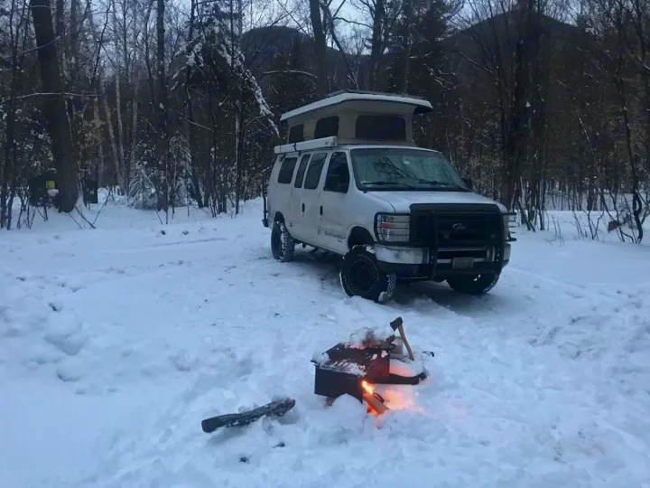 Winter RV rental adventure: Pop-top van in a snowy forest with a crackling campfire. Perfect for cold weather camping.. Ford E250 2008