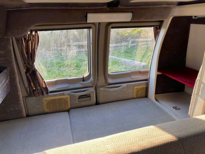 Interior of an RV rental showing a comfy bed, windows with curtains, and built-in storage. Ideal for road trips!. Ford E250 2008