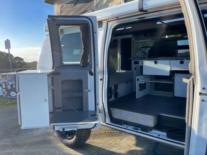 White RV rental interior via open side door. Features a compact kitchen, bed, and external storage unit. Ready for your next adventure.. Ford E250 2008