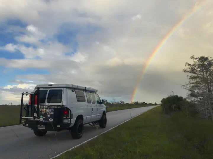 Explore nature in an RV rental! A custom van on a wet road, with a bright rainbow arching over a marshy landscape.. Ford E250 2008