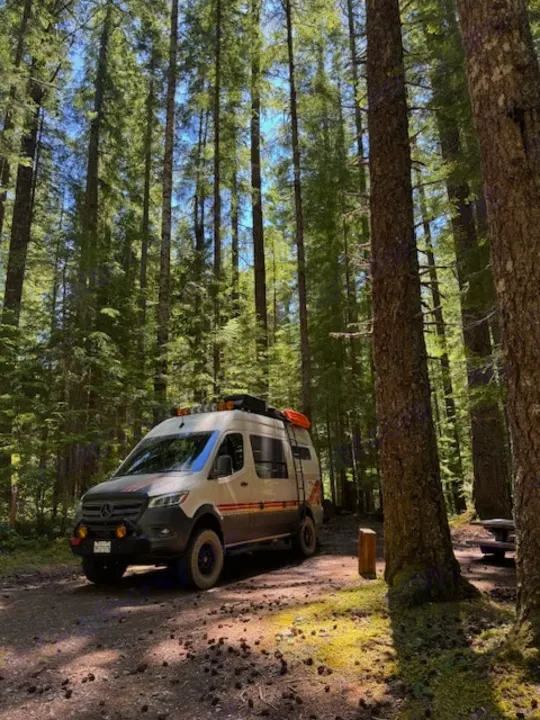 Adventure-ready RV rental in a serene forest campsite. Customized camper van under tall trees, perfect for off-road travel.. Storyteller Overland Storyteller Overland Class B 2023
