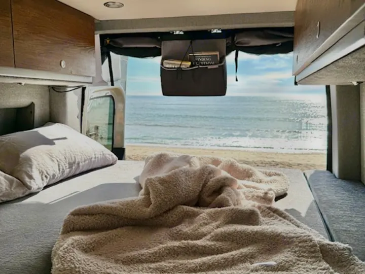 RV rental bedroom: A cozy bed inside a camper van, offering a stunning view of a sunny beach and ocean.. Storyteller Overland Storyteller Overland Class B 2023