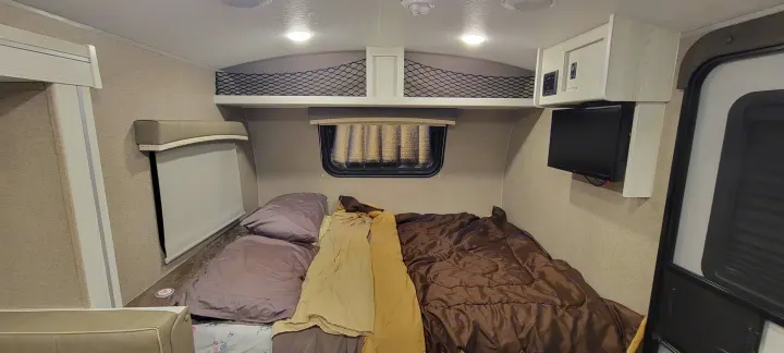 Inside a compact RV rental, showing a neatly made bed with pillows, an overhead storage net, window, TV, and entrance door.. Forest River Rockwood Geo Pro 2022